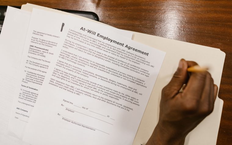 Close-up of a hand signing an at-will employment agreement on a wooden desk. Gros plan d'une main signant un accord d'emploi à volonté sur un bureau en bois.