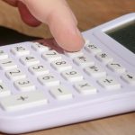 A hand pressing keys on a white calculator placed on a wooden desk in bright daylight. Une main appuyant sur les touches d'une calculatrice blanche posée sur un bureau en bois à la lumière du jour.