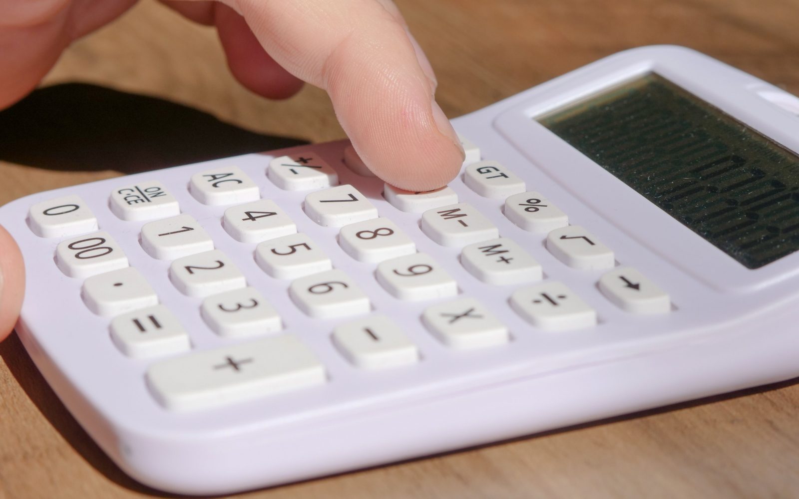 A hand pressing keys on a white calculator placed on a wooden desk in bright daylight. Une main appuyant sur les touches d'une calculatrice blanche posée sur un bureau en bois à la lumière du jour.