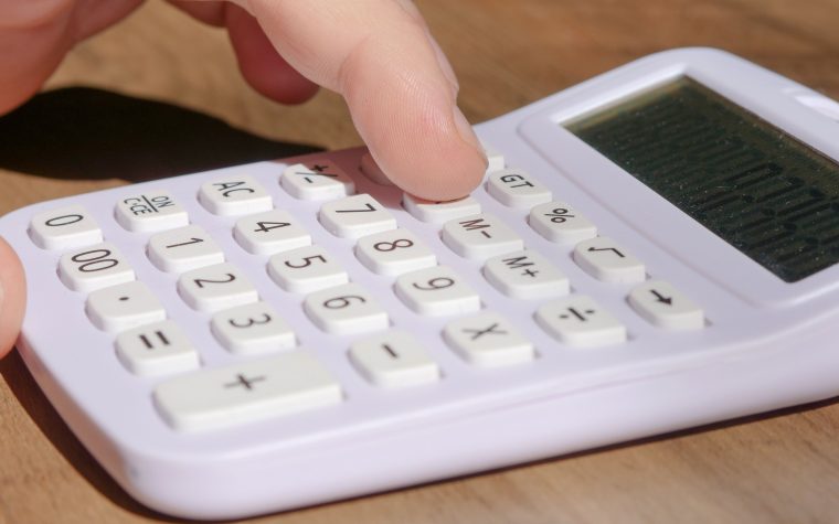 A hand pressing keys on a white calculator placed on a wooden desk in bright daylight. Une main appuyant sur les touches d'une calculatrice blanche posée sur un bureau en bois à la lumière du jour.