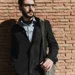 Homme adulte élégant avec barbe et lunettes posant dans une veste noire à l'extérieur contre un mur de briques.
