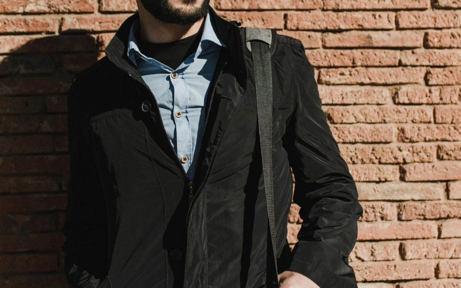 Homme adulte élégant avec barbe et lunettes posant dans une veste noire à l'extérieur contre un mur de briques.