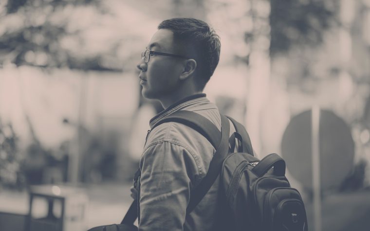 Black and white portrait of a man wearing eyeglasses with a backpack in an outdoor urban setting. Portrait en noir et blanc d'un homme portant des lunettes avec un sac à dos dans un cadre urbain extérieur.