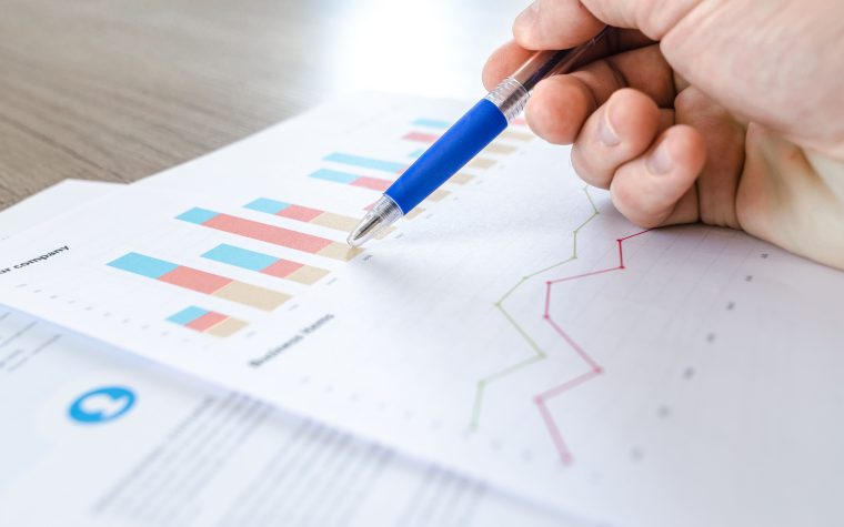 A close-up of a hand with a pen analyzing data on colorful bar and line charts on paper. Gros plan d'une main avec un stylo analysant des données sur des graphiques en barres et en courbes colorés sur papier.